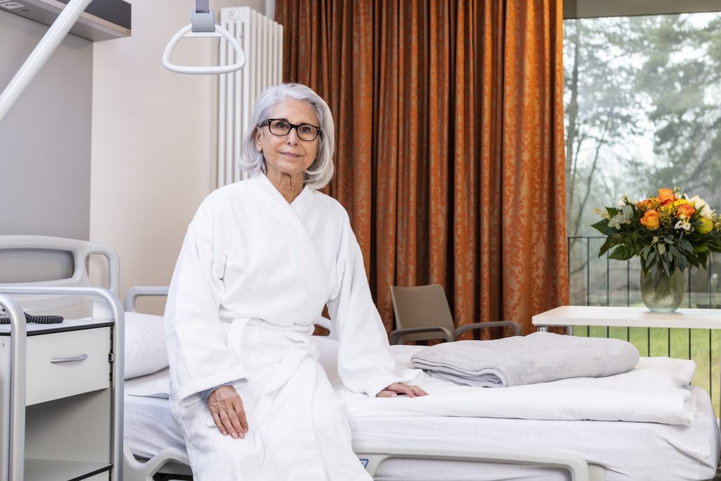 Auf dem Foto sieht man eine Patientin im Bademantel auf dem Patientenbett sitzend. Neben dem Bett steht ein Nachtschrank. Auf dem Bett liegt eine hellgraue Wolldecke. Im Hintergrund erkennt man einen Blumenstrauß, der auf dem Tisch steht, das Fenster, durch welches man die Parkanlage, der Klinik Wartenberg sehen kann, sowie einen orangefarbenen Vorhang.