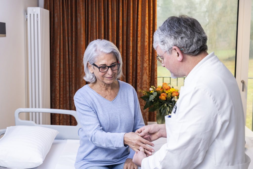 Auf dem Foto sieht man den Chefarzt Doktor Klaus-Friedrich Becher bei der Untersuchung einer Patientin. Die Patientin sitzt auf dem Bett und hält dem Chefarzt ihre Hand entgegen. Der Chefarzt steht mit der Schulter zur Kamera gewandt und untersucht die Hand der Patientin. Im Hintergrund sieht man einen Blumenstrauß auf dem Tisch stehen, das Fenster, durch welches man die begrünte Anlage, der Klinik Wartenberg sehen kann, sowie einen orangenen Vorhang.