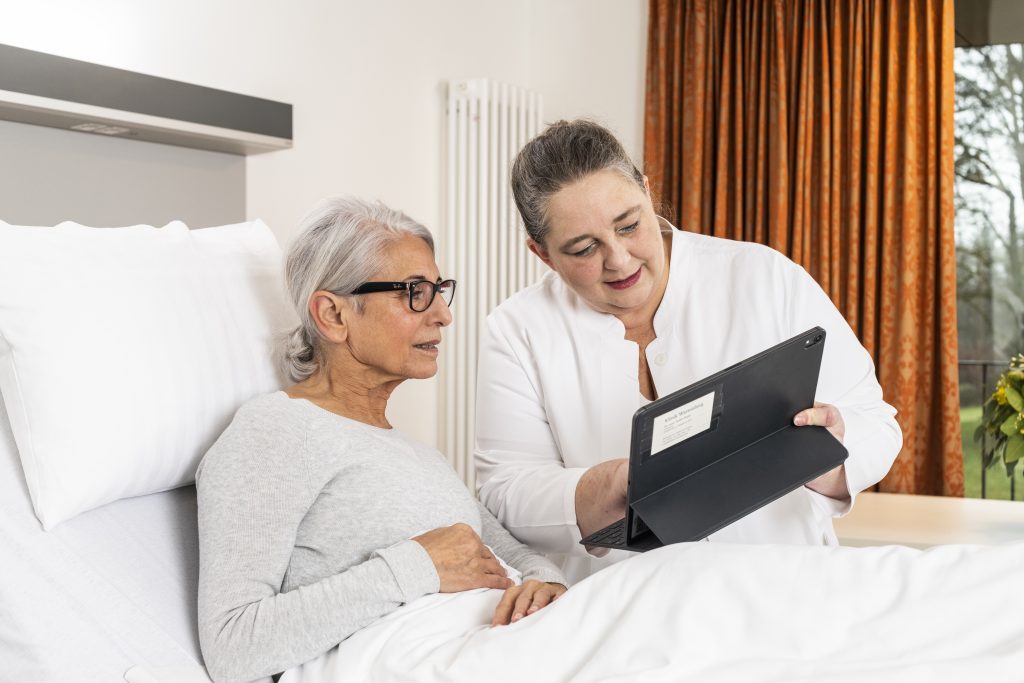 Auf dem Foto sieht man die Chefärztin, Frau Doktor Rupp, wie Sie einer Patientin, die im Bett liegt, auf dem Tablet etwas zeigt. Die beiden Frauen schauen konzentriert auf das Tablet. Im Hintergrund erkennt man das Fenster des Patientenzimmers mit einem orangenen Vorhang.