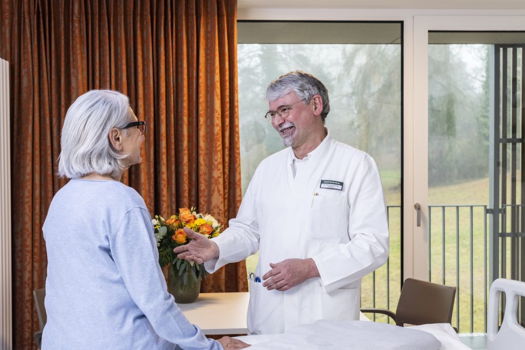 Auf dem Foto sieht man den Chefarzt Doktor Klaus-Friedrich Becher im Patientenzimmer zu einer Patientin gewandt, die auf dem Bett sitzt und ihn anschaut. Die Patientin ist mit der Schulter zur Kamera gewandt. Der Chefarzt hält ihr lächelnd die Hand entgegen. Im Hintergrund sieht man einen Blumenstrauß auf dem Tisch stehen, daneben einen Stuhl, das Fenster, durch welches man die begrünte Anlage, der Klinik Wartenberg sehen kann, sowie einen orangefarbenen Vorhang.