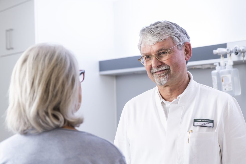 Auf dem Foto sieht man den Chefarzt Doktor Klaus-Friedrich Becher in einem Patientenzimmer einer Patientin gegenüberstehend. Die Patientin sitzt mit dem Rücken zur Kamera gewandt. Im Hintergrund erkennt man das Patientenzimmer.