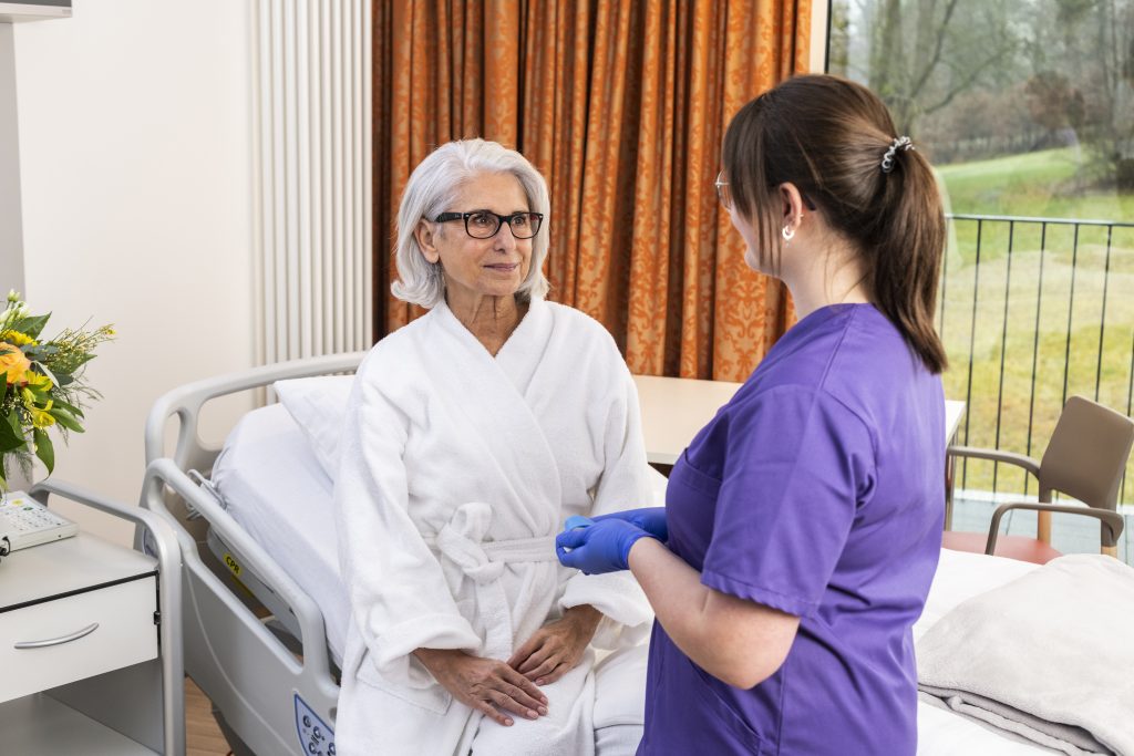 Auf dem Foto sieht man die medizinische Fachangestellte Eleonora Carbone in einem violetten Kasak mit der Schulter zur Kamera gewandt, im Patientenzimmer stehend. Die MFA schaut eine Patientin an, die im Bademantel auf dem Patientenbett sitzt und zur Fachkraft schaut. Man sieht den Nachtschrank mit einem Blumenstrauß darauf, im Hintergrund einen Tisch mit Stuhl vor dem Fenster, neben dem ein orangener Vorhang hängt.
