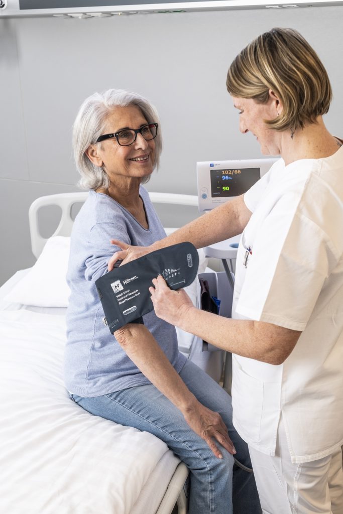 Auf dem Foto sieht man die Stationsleitung Jane Hofmayr, wie sie einer Patientin, die auf dem Bett sitzt, die Manschette zur Blutdruckmessung anlegt. Die Patientin schaut zur Pflegekraft und lächelt sie an. Im Hintergrund sieht man das Gerät zur Vitalzeichenmessung.