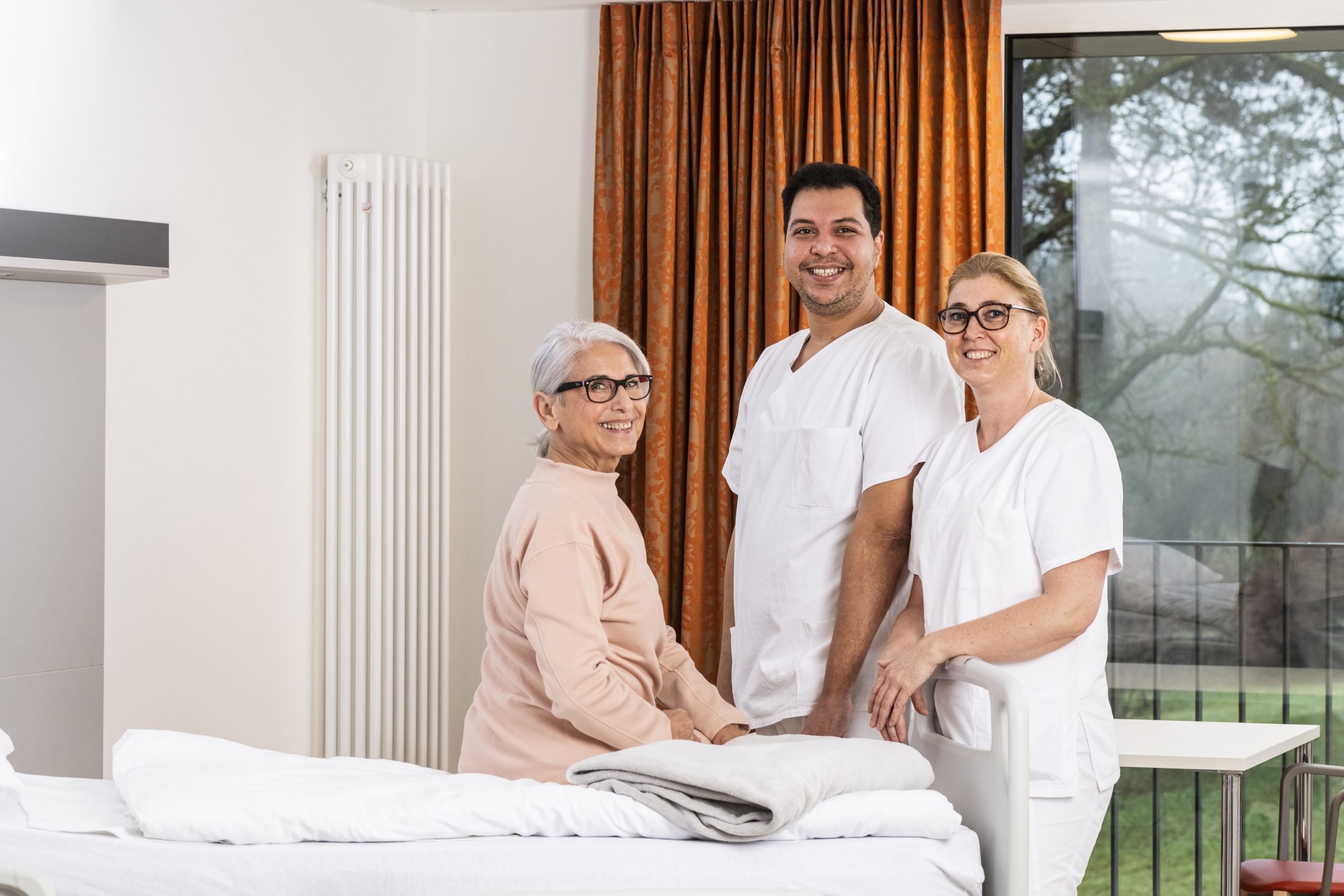 Auf dem Foto sieht man zwei Pflegefachkräfte zusammen mit einer Patientin, die auf dem Bett sitzt. Die drei Personen stehen im Patientenzimmer und lächeln in die Kamera. Im Hintergrund sieht man das Fenster, durch welches man in den begrünten Innenhof schauen kann. Neben dem Fenster hängt ein orangener Vorhang.