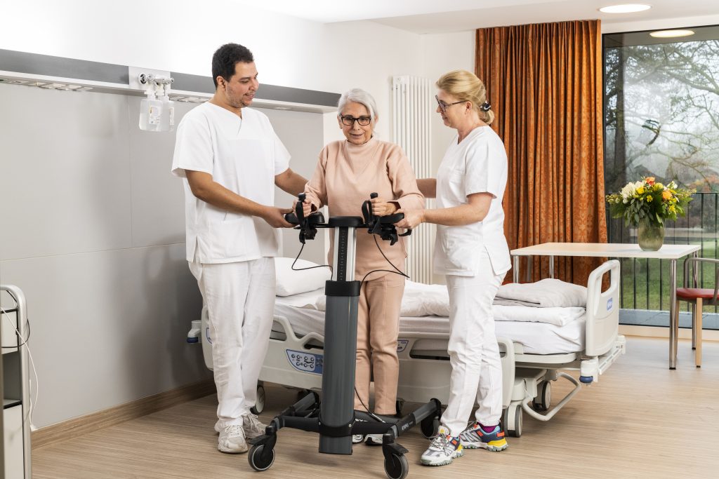 Auf dem Foto sieht man zwei Pflegefachkräfte, die eine Patientin im beigefarbenen Trainingsanzug an einem Gerät unterstützen. Die Patientin hält sich mit beiden Händen an dem Gerät fest, und die Pflegefachkräfte stützen Sie von rechts und links. Die drei Personen stehen im Patientenzimmer, im Hintergrund sieht man das Patientenbett, einen Tisch mit Blumenstrauß und das Fenster, durch das man in den Innenhof schauen kann. Neben dem Fenster hängt ein orangener Vorhang.