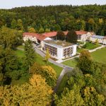 Das Foto zeigt eine Luftaufnahme, der Klinik Wartenberg von Südwesten aus. Mittig ist der Neubau der Klinik zu sehen und dahinter erstreckt sich das weitere Gebäude. Links im Bild erkennt man das Haus Irmgard. Die Klinik ist umgeben von einem grünen Park- und Waldareal.