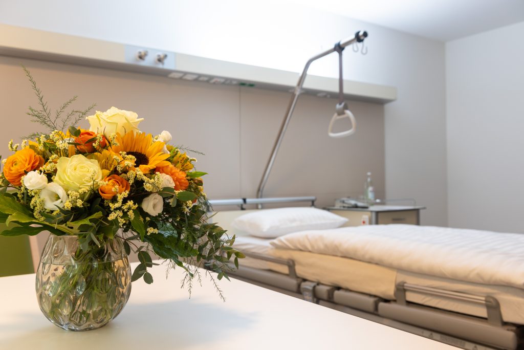 Das Foto zeigt einen Ausschnitt eines Patientenzimmers im Neubau der Klinik Wartenberg. Im Vordergrund steht ein Blumenstrauß auf dem Tisch. Dahinter erkennt man einen Patientenbett mit einem Nachtschrank.