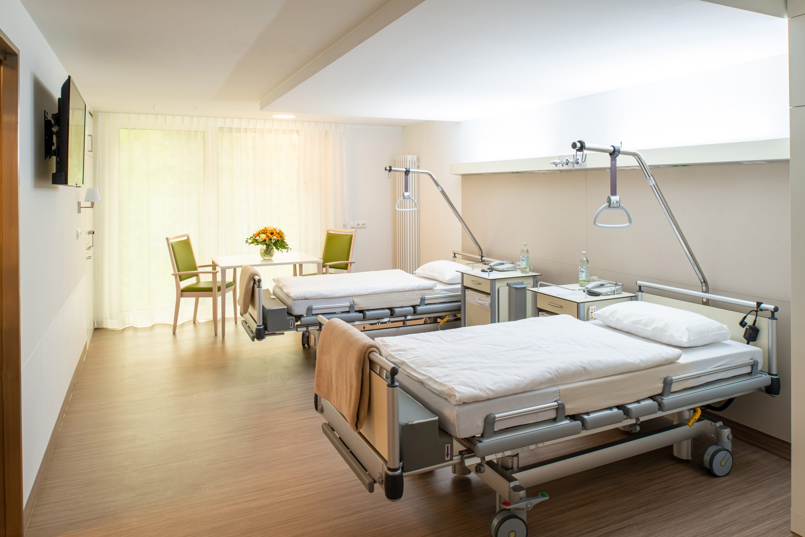 Auf dem Foto sieht man ein Doppelzimmer im Neubau der Klinik Wartenberg. Darin stehen zwei Betten mit je einem Nachtschrank mit Telefon und Flasche Wasser darauf. Im Hintergrund steht ein Tisch mit zwei Stühlen und einem Blumenstrauß. Links an der Wand hängt ein Fernseher. Im Hintergrund sieht man die großen Fenster, durch welche man die begrünte Parkanlage der Klinik Wartenberg sehen kann.