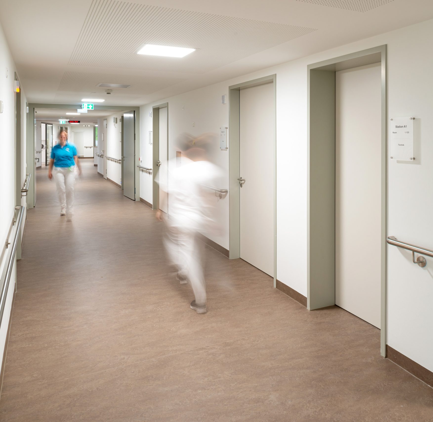 Auf dem Foto sieht man den Stationsflur einer akutgeriatrischen Station im Neubau der Klinik Wartenberg. Eine Pflegekraft sowie eine Patientin Betreuerin gehen über den Flur.