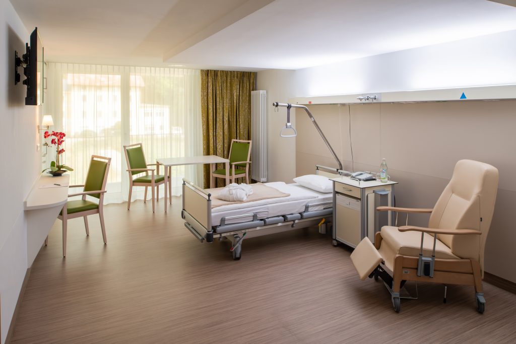 Auf dem Foto sieht man ein modernes Patientenzimmer im Neubau der Klinik Wartenberg. Rechts im Bild steht ein beigefarbener Sessel. In der Mitte des Raumes steht ein Patientenbett mit beigefarbener Wolldecke und einem weißen, gefalteten Bademantel darauf. Daneben steht ein Nachtschrank mit Telefon und einer Flasche Wasser darauf. Vor der großen Fensterfront steht ein Tisch mit zwei Stühlen. Links im Bild befindet sich ein weiterer Stuhl vor einem kleinen Schreibtisch. Darüber hängt ein Fernseher an der Wand. Im Hintergrund sieht man die Fenster mit dem Balkon, der zum Innenhof, der Klinik Wartenberg raus zeigt.
