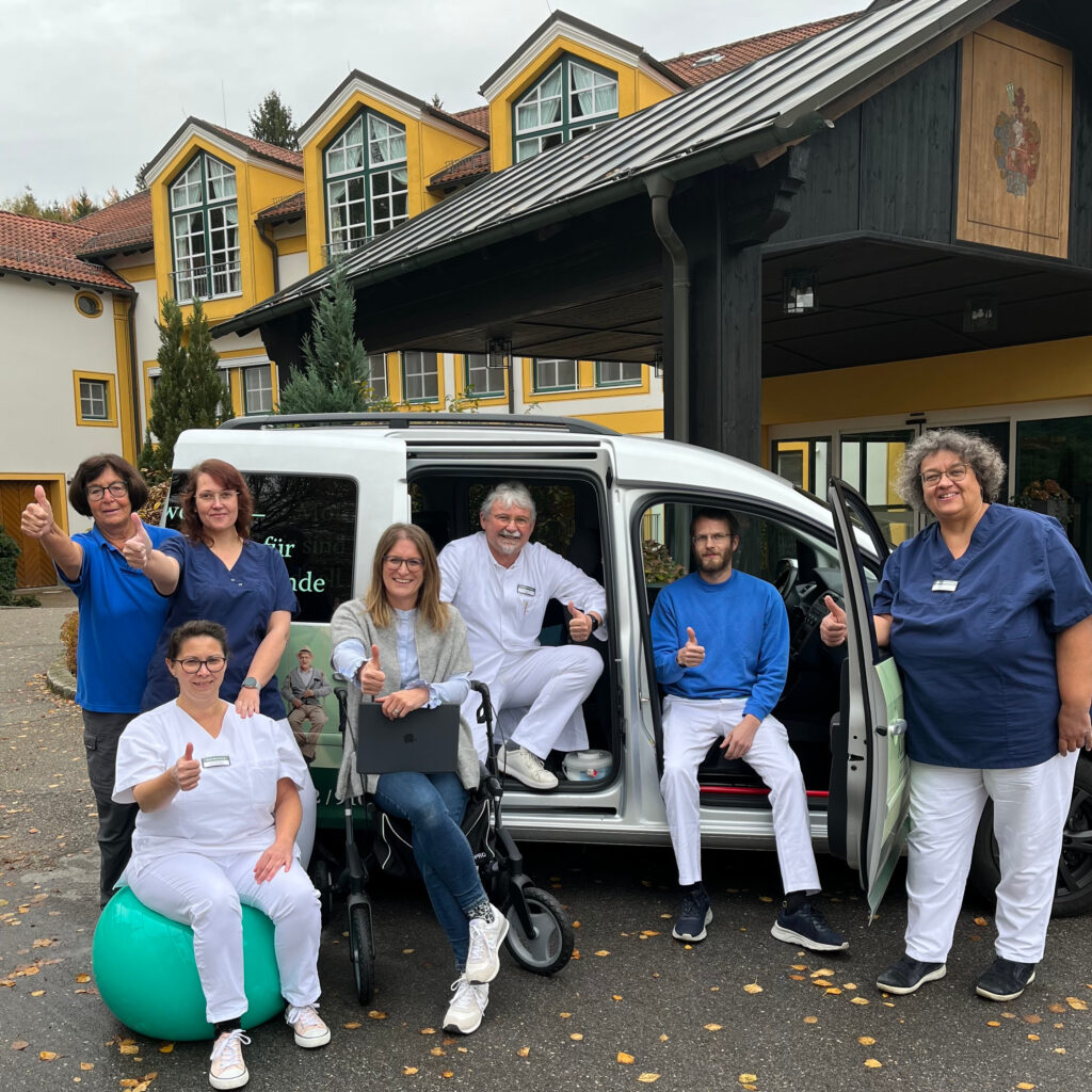 Foto der Mitarbeiterinnen und Mitarbeiter, die für die Mobile geriatrische Reha unterwegs sind. Die Mitarbeitenden sitzen und stehen um ein Fahrzeug der MoGeRe der Klinik Wartenberg herum und halten einen Daumen hoch in die Kamera. Das Fahrzeug steht vor dem Eingangsbereich der Klinik Wartenberg.
