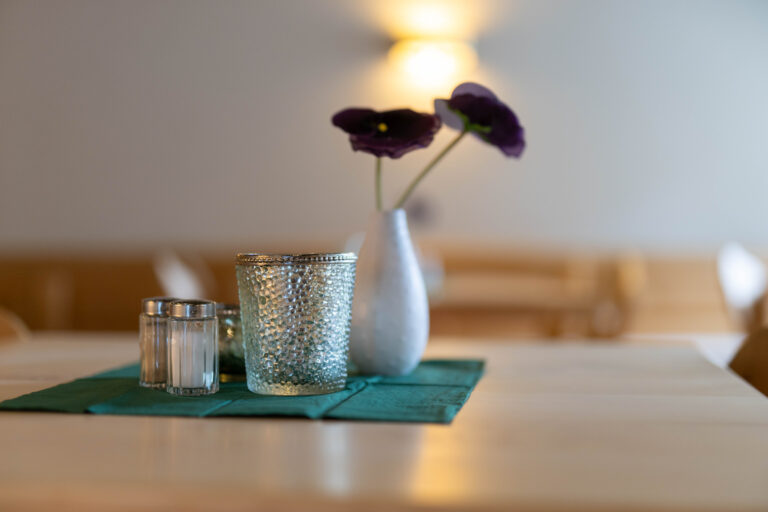 Auf dem Foto sieht man einen Tisch im Bistro der Klinik Wartenberg. Darauf stehen Salz- und Pfefferstreuer, Teelichter und eine kleine Vase mit einer dunkelvioletten Blume darin auf einer grünen Serviette. Im Hintergrund erkennt man unscharf weitere Tische. Eine Lampe leuchtet an der Wand.