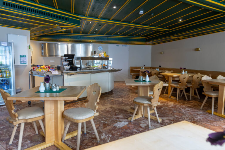 Auf dem Foto sieht man das Bistro der Klinik Wartenberg. Im Raum verteilt stehen Tische mit Stühlen. Auf den Tischen befinden sich kleine Vasen, Teelichter und Salz- und Pfefferstreuer auf grünen Servietten. Im Hintergrund sieht man die Essensausgabe mit der Kasse und der Auslage. Dahinter sind silberne Schränke zu erkennen. Die Decke ist Dunkelgrün und gelb. Links im Bild sieht man einen Kühlschrank mit Getränken.