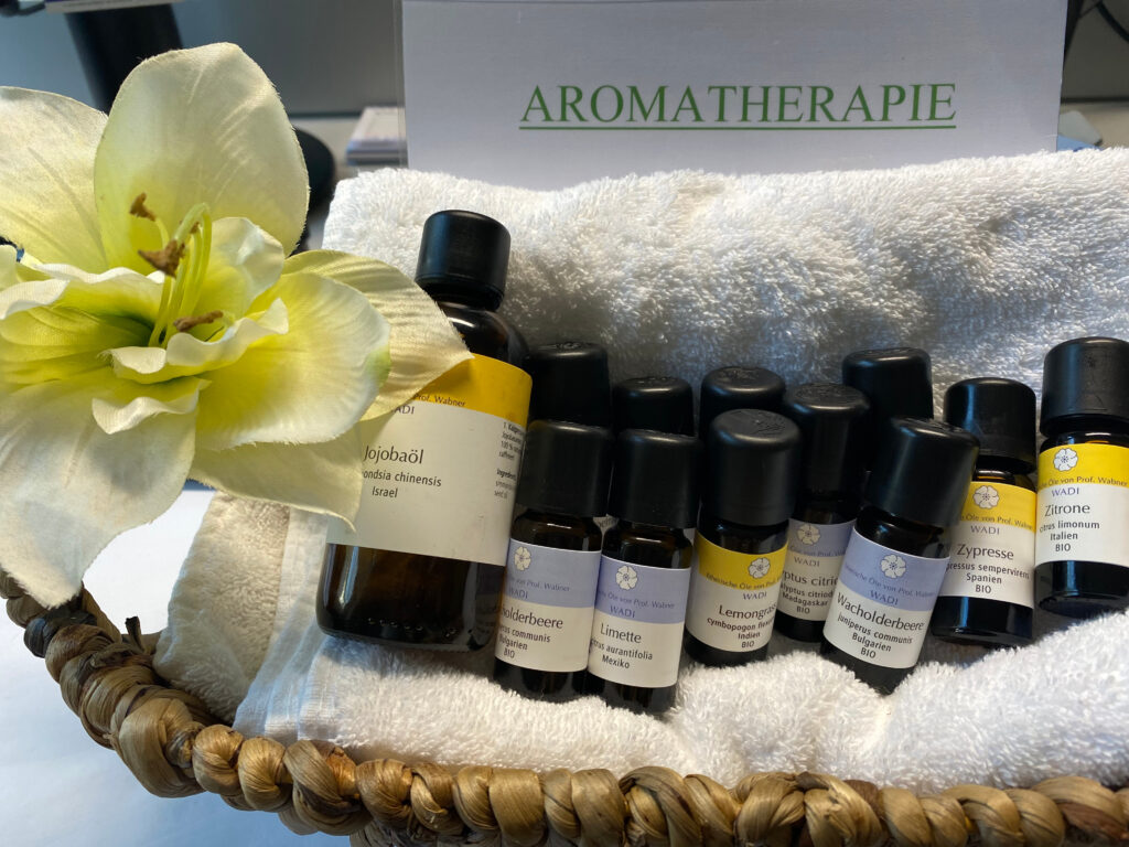 Foto eines Körbchens mit verschiedenen ätherischen Ölen, die auf einem Handtuch liegen. Links im Bild sieht man eine Blumenblüte und im Hintergrund steht die Aufschrift 'Aromatherapie'.