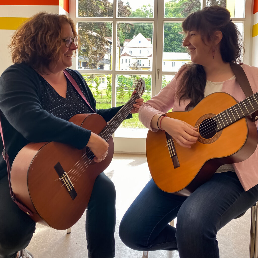 Foto von zwei Frauen, die Gitarre spielen und sich dabei lächelnd anschauen. Die beiden Frauen sitzen im Gymnastiksaal der Klinik Wartenberg und durch das Fenster im Hintergrund kann man in den Innenhof schauen.