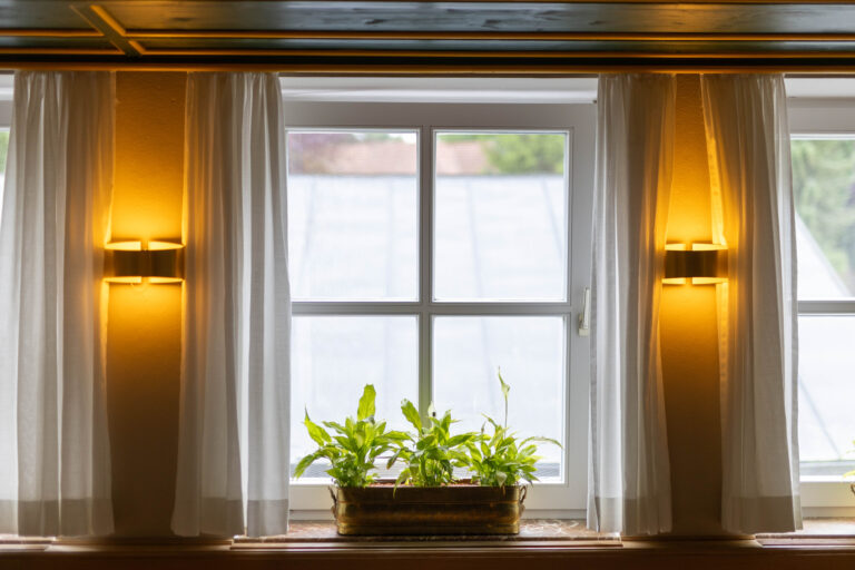 Ein Foto der Fenster im Bistro der Klinik Wartenberg. In der Mitte auf der Fensterbank stehteine grüne Pflanze in eine, goldenen Topf. Zwischen den Fenstern leuchten jeweils Lampen und rechts und links davon hängen weiße Vorhänge. Durch die Fenster sieht man das Dach des Eingangsbereichs der Klinik.