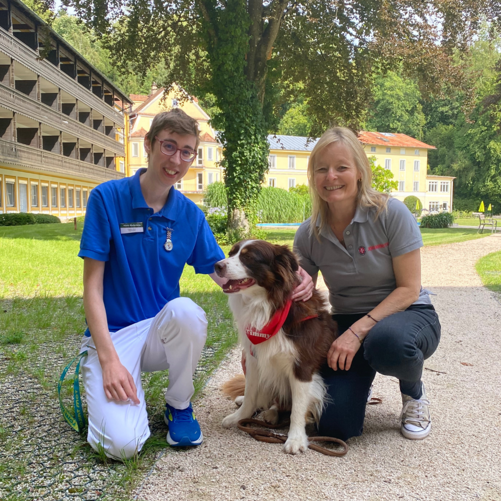 Foto einer Ergotherapeutin zusammen mit dem Therapiehund Timmy und seiner Besitzerin von den Johannitern, die zusammen im Innenhof der Klinik Wartenberg sitzen.