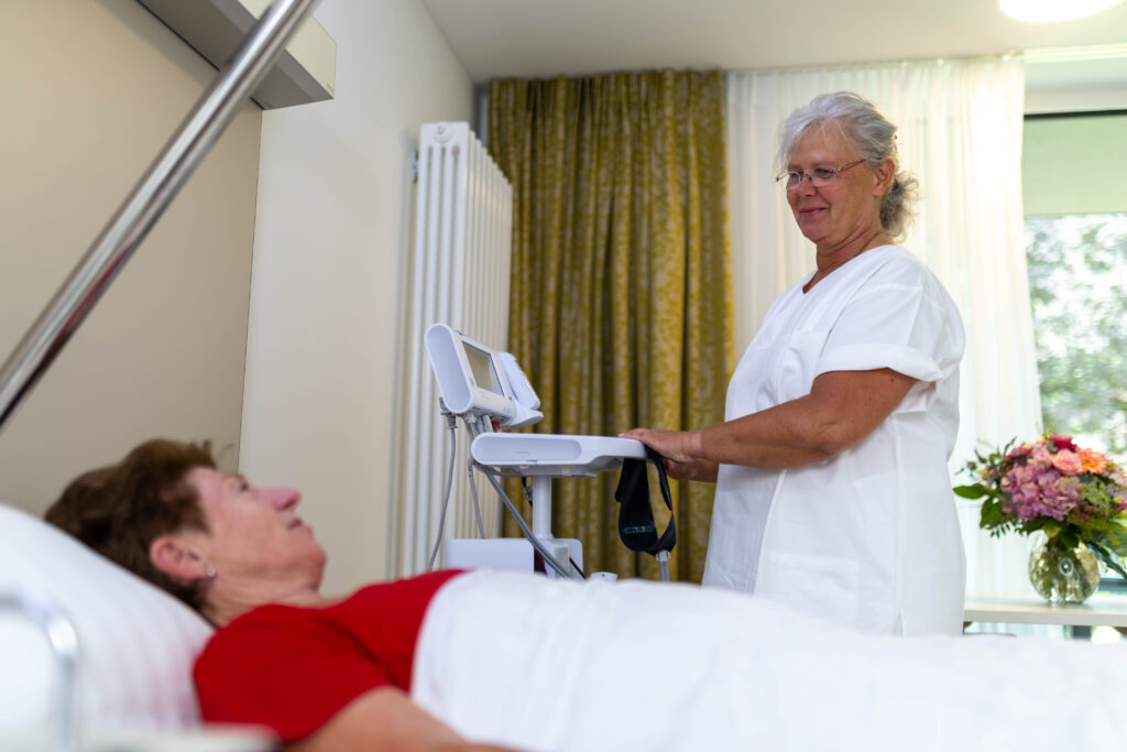 Auf dem Foto sieht man die Pflegefachkraft Ronny Schoß im Patientenzimmer neben einer Patientin, die im Bett liegt. Die beiden Frauen lächeln einander an. Die Pflegefachkraft hält das Gerät zur Vitalzeichenmessung fest. Im Hintergrund sieht man einen Blumenstrauß auf dem Tisch stehen sowie das Fenster durch das man in den Innenhof schauen kann.