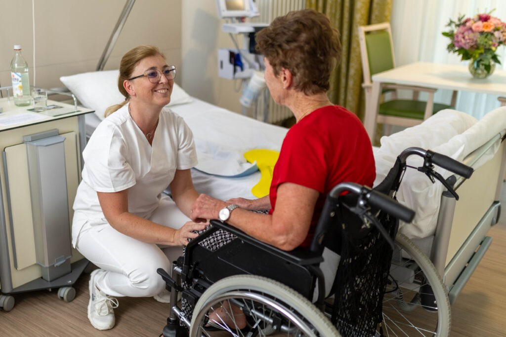 Auf dem Foto sieht man die Pflegefachkraft Susanne Leise in der Hocke vor einer Patientin, die im Rollstuhl im Patientenzimmer sitzt. Die Pflegefachkraft lächelt die Patientin an, welche seitlich mit dem Rücken zur Kamera gewandt sitzt. Im Hintergrund erkennt man das Patientenbett, Nachtschrank, Stuhl und Tisch mit einem Blumenstrauß darauf, sowie das Messgerät zur Vitalzeichenmessung.