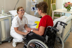Auf dem Foto sieht man die Pflegefachkraft Susanne Leise in der Hocke vor einer Patientin, die im Rollstuhl im Patientenzimmer sitzt. Die Pflegefachkraft lächelt die Patientin an, welche seitlich mit dem Rücken zur Kamera gewandt sitzt. Im Hintergrund erkennt man das Patientenbett, Nachtschrank, Stuhl und Tisch mit einem Blumenstrauß darauf, sowie das Messgerät zur Vitalzeichenmessung.