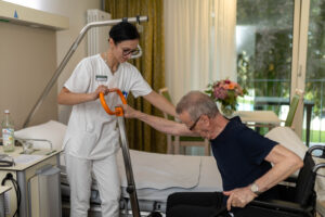 Auf dem Foto sieht man eine Pflegefachkraft im Patientenzimmer, wie sie einem Patienten hilft, mithilfe eines Hilfsgeräts aus dem Rollstuhl aufzustehen. Der Patient hält sich mit einer Hand an dem Hilfsgerät fest und lehnt sich vor. Die Pflegefachkraft hält ihn am Arm fest und unterstützt ihn beim Aufstehen. Im Hintergrund sieht man das Patientenbett sowie den Tisch im Patientenzimmer. Hinten rechts im Bild erkennt man das Fenster und den Balkon, hinter dem sich der Innenhof befindet.