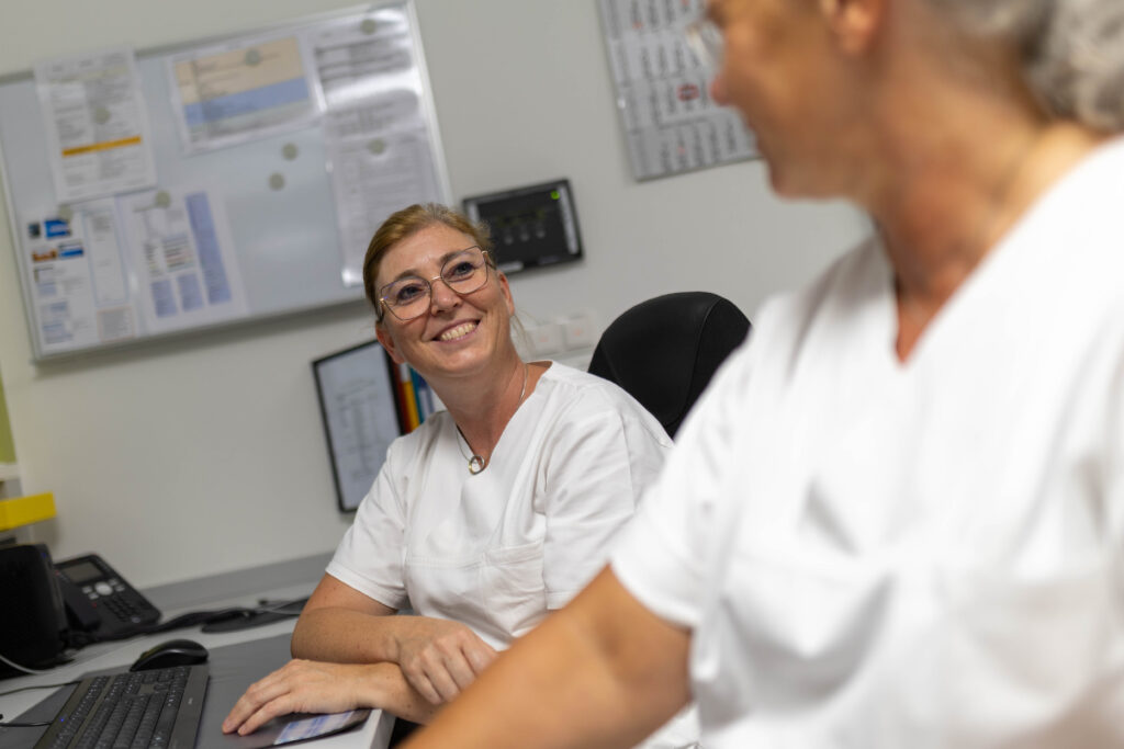 Das Foto zeigt die Stationsleitung Susanne Leise zusammen mit Ronny Schoß im Pflegestützpunkt der akutgeriatrischen Station. Susanne schaut Ronny an, die unscharf im Vordergrund zu erkennen ist. Beide sitzen am Schreibtisch vor ihren Arbeitsplätzen. Im Hintergrund erkennt man ein Whiteboard mit diversen Dokumenten darauf sowie einen Kalender.