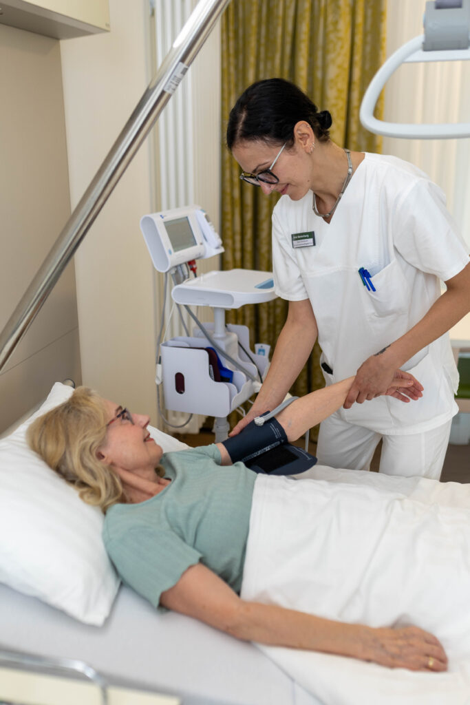 Eine Pflegefachkraft misst den Blutdruck einer Patientin, die im Bett im Patientenzimmer liegt. Die Pflegekraft legt ihr die Blutdruck-Manschette um den linken Arm und hält diesen mit der Hand fest. Neben ihr steht das Gerät zur Vitalzeichenmessung. Im Hintergrund sieht man den Vorhang vor dem Fenster des Patientenzimmers.