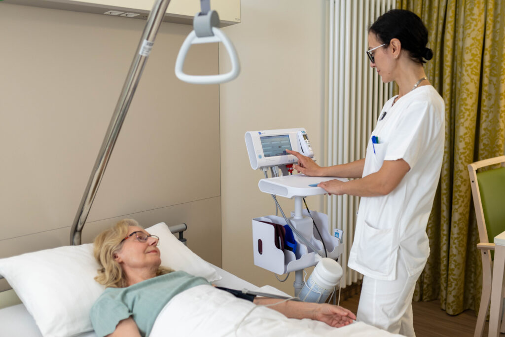 Eine Pflegefachkraft steht neben einer Patientin, die im Bett liegt, im Patientenzimmer und misst mithilfe des Vitalzeichen-Messgerätes ihren Blutdruck. Im Hintergrund sieht man die Heizung, einen Vorhang und einen Stuhl.