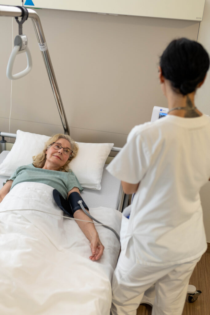 Eine Pflegekraft steht mit dem Rücken zur Kamera gewand im Patientenzimmer. Die Patientin liegt im Bett und trägt eine Blutdruck-Manschette um ihren linken Arm. Die Pflegekraft schaut auf das Gerät zur Vitalzeichenmessung, welches neben dem Patientenbett steht.