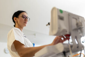 Eine Pflegefachkraft im weißen Kasak schaut auf das Gerät zur Vitalzeichenmessung, welches im Vordergrund des Bildes unscharf zu erkennen ist. Im Hintergrund erkennt man ein Patientenzimmer.