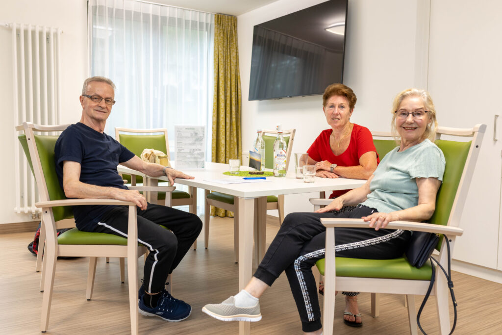 Ein Patient und zwei Patientinnen sitzen am Tisch im Aufenthaltsraum einer akutgeriatrischen Station. Sie schauen zur Kamera. Auf dem Tisch stehen Wasserflaschen und Gläser. Im Hintergrund sieht man einen Fernseher an der Wand sowie ein Fenster mit einem durchscheinen Vorhang davor.
