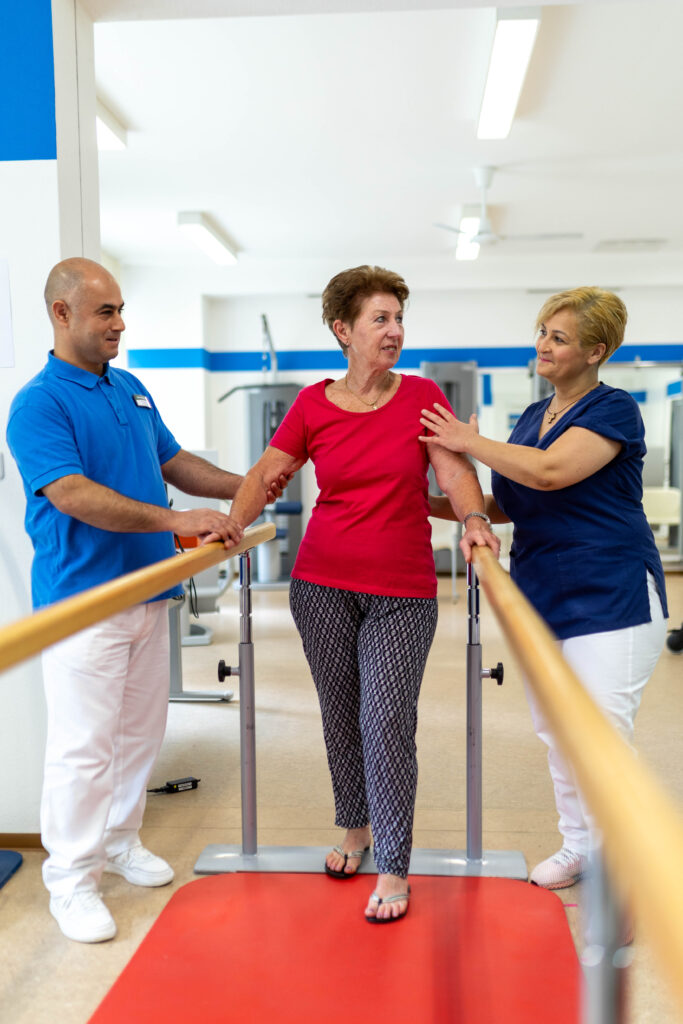 Zwei Physiotherapeuten unterstützen eine Patienten beim gehen am Geh-Barren. Die Patientin steht mittig zwischen den Griffstangen des Geräts und die Therapeuten stehen rechts und links daneben und halten die Patientin an den Armen. Sie steht auf deiner roten Sportmatte. Im Hintergrund erkennt man die Geräte im MTT-Raum.
