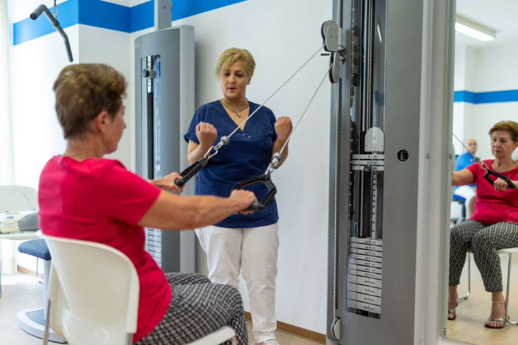 Eine Physiotherapeutin leitet eine Patientin beim Training mit dem Seilzug an. Die Patienten sitzt auf einem Stuhl seitlich von der Kamera abgewandt und zieht die Griffe des Seilzugs seitlich nach unten. Die Therapeutin zeigt ihr, wie sie die Übung durchführen soll. Im Spiegel rechts im Bild sieht man die Spiegelung der Patientin sowie eines weiteren Physiotherapeuten im Hintergrund. Im Hintergrund steht ein weiteres Trainingsgerät im MTT-Raum der Klinik Wartenberg.