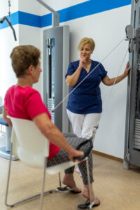 Eine Physiotherapeutin beobachtet eine Patientin beim Training am Seilzug. Die Patienten sitzt auf einem Stuhl seitlich von der Kamera abgewandt und zieht die Griffe des Seilzugs seitlich nach unten. Die Physiotherapeutin steht neben dem Gerät und schaut der Patientin beim Training zu. Im Hintergrund steht ein weiteres Trainingsgerät im MTT-Raum der Klinik Wartenberg.