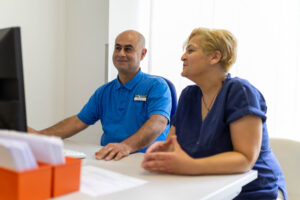Foto von zwei Physiotherapeuten am Arbeitsplatz. Beide tragen blaue Arbeitskledidung. Sie sitzen am Schreibtisch und schauen auf den Monitor.