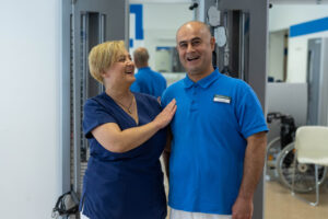 Foto von zwei Physiotherapeuten im MTT-Raum der Klinik Wartenberg. Sie halten einander im Arm und lachen fröhlich. Beide tragen die blaue Arbeitskleidung. Im Hintergrund sieht man Sportgeräte und Spiegel.