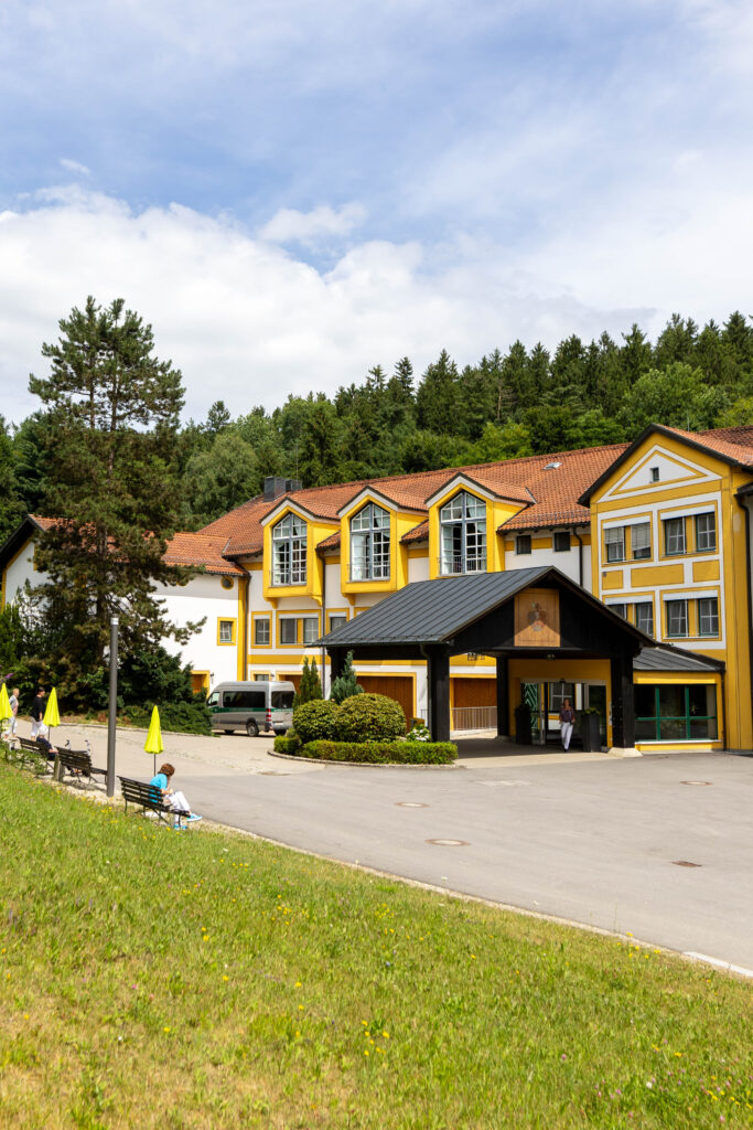 Foto des Haupteingangs der Klinik Wartenberg. Der Eingangsbereich ist vorgelagert und überdacht. Dahinter befindet sich der Bestandsbau, der gelb-weiß gestrichen ist. Im oberen Teil des Gebäudes erkennt man die großen Fenster des Vortragssaals. Vor dem Gebäude ist die asphaltierte Zufahrt zum Eingang. Im Vordergrund des Bildes sieht man die grüne Wiese, die sich gegenüber vom Eingang befindet. Am Rand stehen Bänke und Sonnenschirme. Der Himmel ist blau und leicht bewölkt. Es scheint ein sommerlicher Tag zu sein.