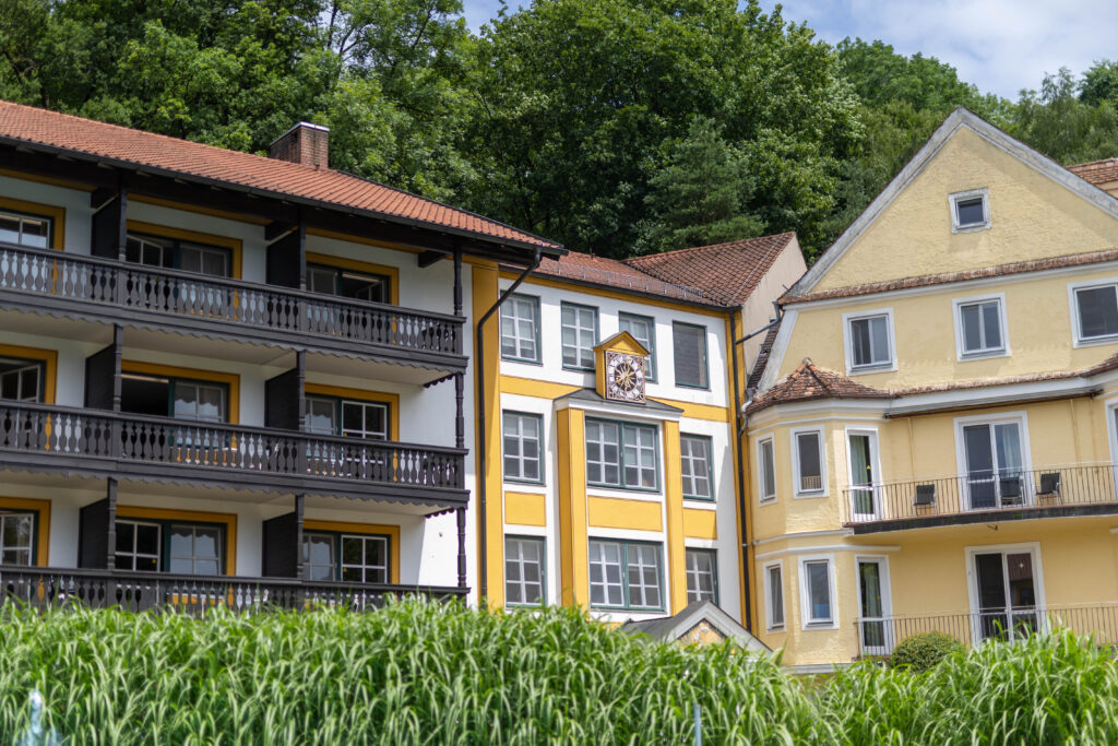 Foto des Klinikgebäudes vom Innenhof der Klinik. Mittig sieht man den Gebäudeübergang von Altbau (rechts) zum Bestandsbau (links). In der Mitte befindet sich eine Uhr an dem weiß-gelben Gebäude. Links sieht man die hölzernen Balkone der Patientenzimmer der Rehabilitations-Stationen. Im Vordergrund sieht man die grünen Pflanzen im Innenhof und im Hintergrund die Bäume des Klinikwaldes.