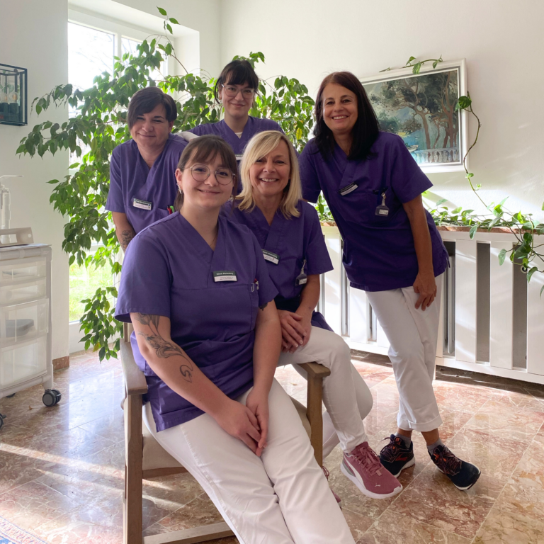 Foto des Teams des medizinischen Funktionsdienstes der Klinik Wartenberg in ihren lilanen Kasacks. Die Gruppe sitzt und steht gemeinsam im Eingangsbereich des Bestandsbaus. Im Hintergrund erkennt man verschiedene Pflanzen, und die Sonne scheint durch das Fenster hinein.