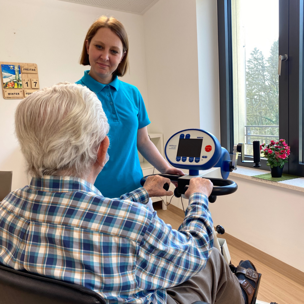 Foto einer Mitarbeiterin der Patientenbetreuung, die einen Patienten bei der Nutzung des E-Radls unterstützt. Die beiden befinden sich im Aufenthaltsraum einer akut geriatrischen Station.