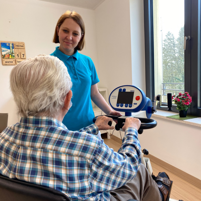 Foto einer Mitarbeiterin der Patientenbetreuung, die einen Patienten bei der Nutzung des E-Radls unterstützt. Die beiden befinden sich im Aufenthaltsraum einer akut geriatrischen Station.