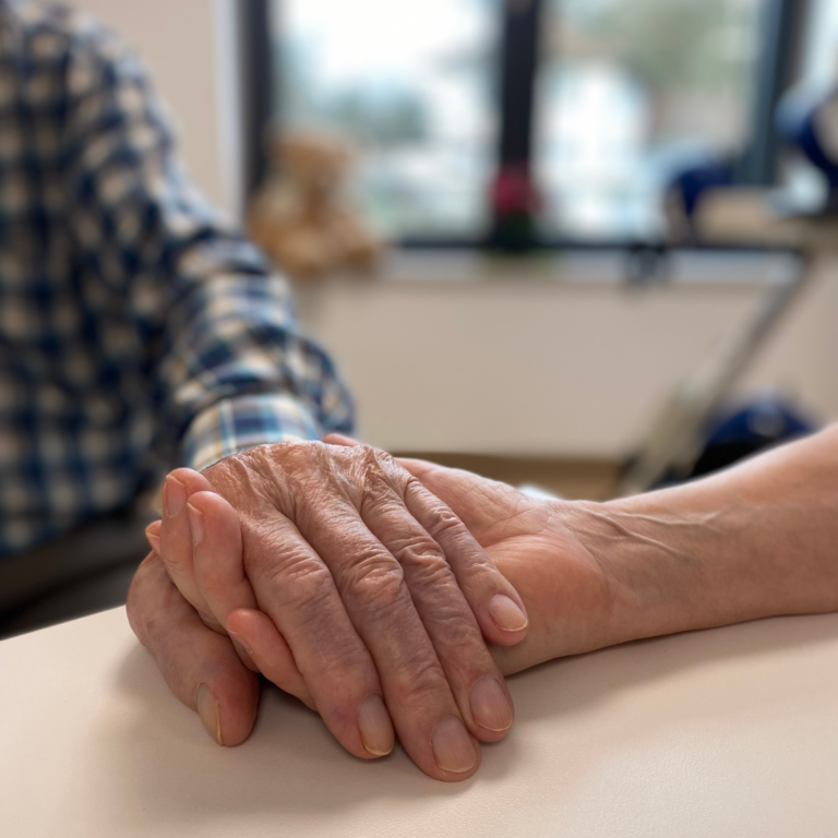 Auf dem Foto sieht man zwei Hände. Die untere Hand einer Frau hält die obere Hand eines älteren Patienten, den man teilweise unscharf im Hintergrund erkennen kann. Im Hintergrund sieht man außerdem ein Fenster, durch das man in den Innenhof der Klinik Wartenberg schauen kann.
