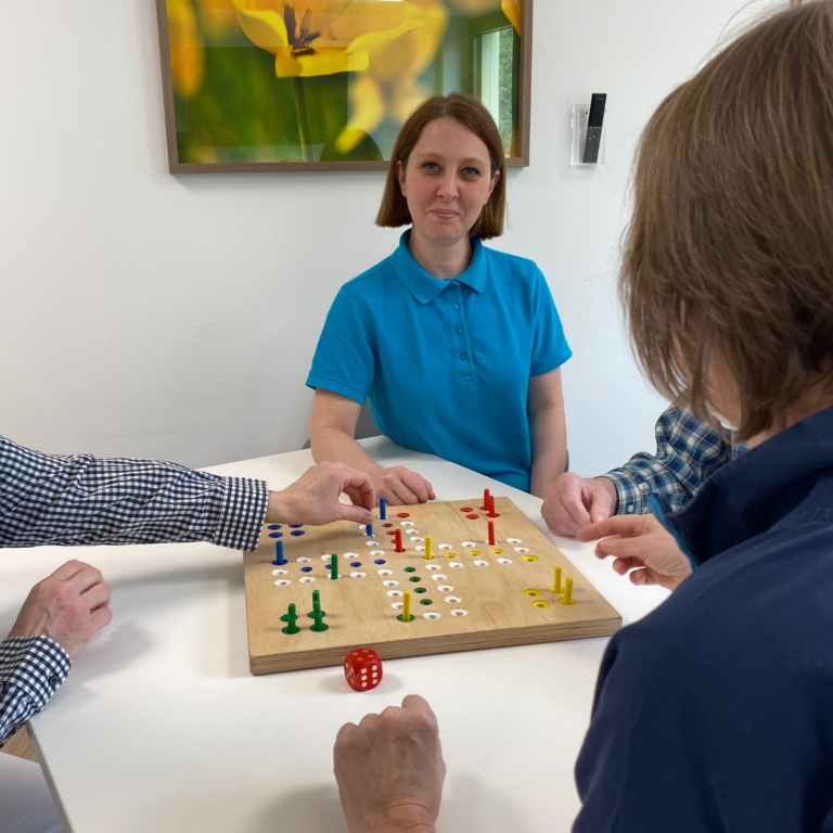 Foto einer Mitarbeiterin der Patientenbetreuung, in ihrem typischen türkisen Poloshirt, die mit mehreren Patient/innen gemeinsam auf einem Tisch Mensch ärgere dich nicht spielt.