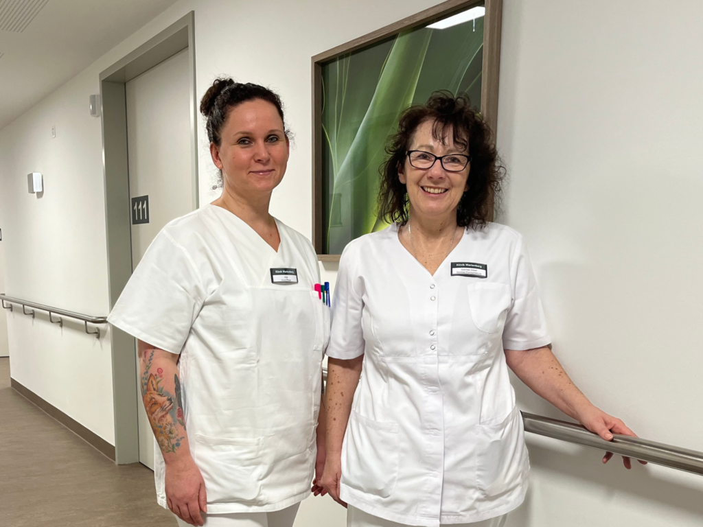 Foto von zwei Pflegefachkräften, die auf dem Stationsflur eine akutgeriatrie Station im Neubau der Klinik Wartenberg stehen und in die Kamera lächeln.