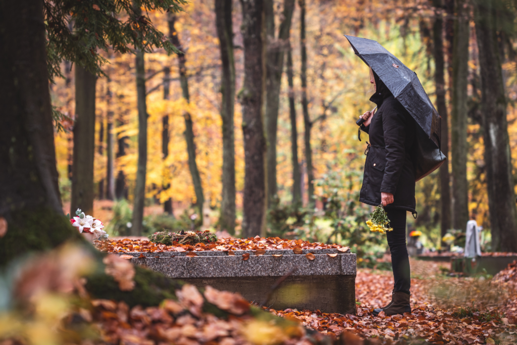 Foto einer Person mit Regenschirm, die an einem Grab steht und einen Blumenstrauß in der Hand hält. Der Friedhof befindet sich in einem Wald, Laub liegt am Boden und die Blätter der Bäume sind gelb-orange gefärbt. Es scheint ein herbstlicher, regnerischer Tag zu sein.