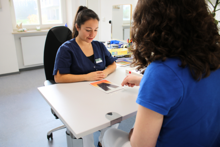 Foto einer Sprachtherapeutin, die mit einer Patientin am Schreibtisch sitzt. Die Patientin legt aus einzelnen Buchstaben das passende Wort zu einer Bildkarte und die Therapeutin beobachtet sie dabei. Im Hintergrund erkennt man weitere Hilfsmittel und Utensilien der Sprachtherapie.