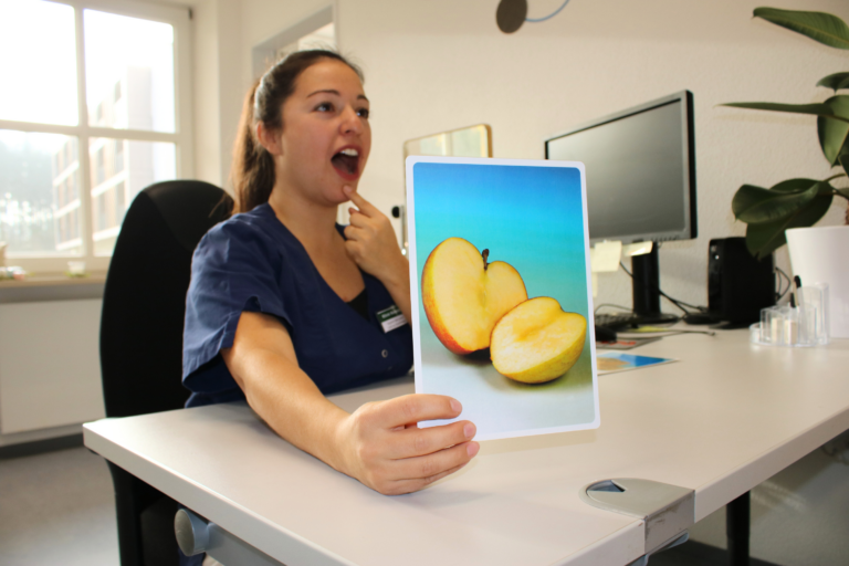 Foto einer Sprachtherapeutin, die eine Bildkarte mit einem Apfel in die Kamera hält und mit der anderen Hand auf ihr Kind zeigt und den Mund geöffnet hat. Sie sitzt an einem Schreibtisch.