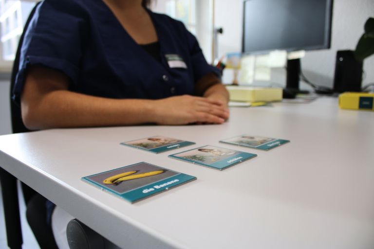 Nahaufnahme der Bildkarten, die in der Sprachtherapie genutzt werden. Im Hintergrund erkennt man die Sprachtherapeutin am Schreibtisch sitzend.