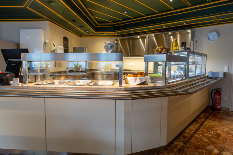 Das Foto zeigt die Essensausgabe im Bistro der Klinik Wartenberg. Man sieht die Auslage für die Speisen mit der Tablettablage. Im Hintergrund erkennt man silberne Schränke. Die Decke ist Dunkelgrün und gelb. Links im Bild befindet sich die Kasse.