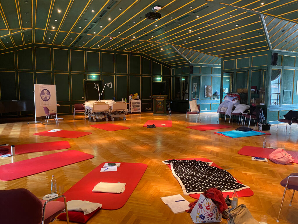 Foto des Gymnastiksaals der Klinik Wartenberg, in dem mehrere Gymnastikmatten auf dem Boden liegen. Im Hintergrund erkennt man 2 Pflegebetten sowie weitere Utensilien. Es handelt sich dabei um eine Fortbildung zur Kinästhetik und basalen Stimulation.